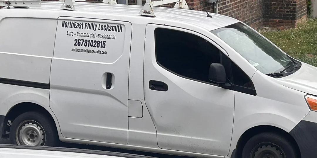 An inconspicuous white van brandishing the name NorthEast Philly Locksmith on the side parked in front of a brick house with green grass poking behind. Reads 'Auto Commercial Residential', with a phone number 2678142815, and the url 'northeastphillylocksmith.com'.